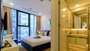 Ein modernes Hotelzimmer mit einem großen Bett, einem Nachttisch, einem Schreibtisch, einem großen Fenster mit Vorhängen sowie einem Badezimmer mit Spiegel und Waschbecken.