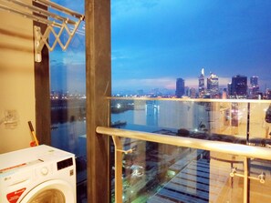 Une pièce équipée d’un lave-linge, offrant une vue sur les toits d’une ville au crépuscule, et un balcon avec une rambarde en verre donnant sur une étendue d’eau.