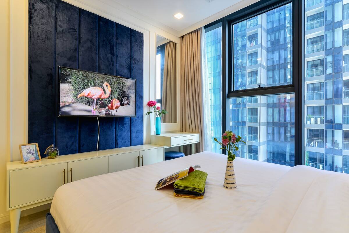 Ein Hotelzimmer mit einem großen Bett, einem flachbildfernseher an einer dunkelblauen Wand, einem Fenster mit Blick auf ein Gebäude und einer Vase mit Blumen auf dem Nachttisch.