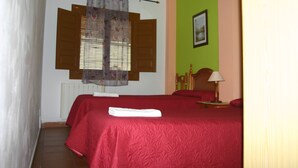 Una habitación con dos camas, una ventana con contraventanas, un radiador, un cabecero de madera, una mesita de noche con una lámpara y un cuadro enmarcado en la pared.