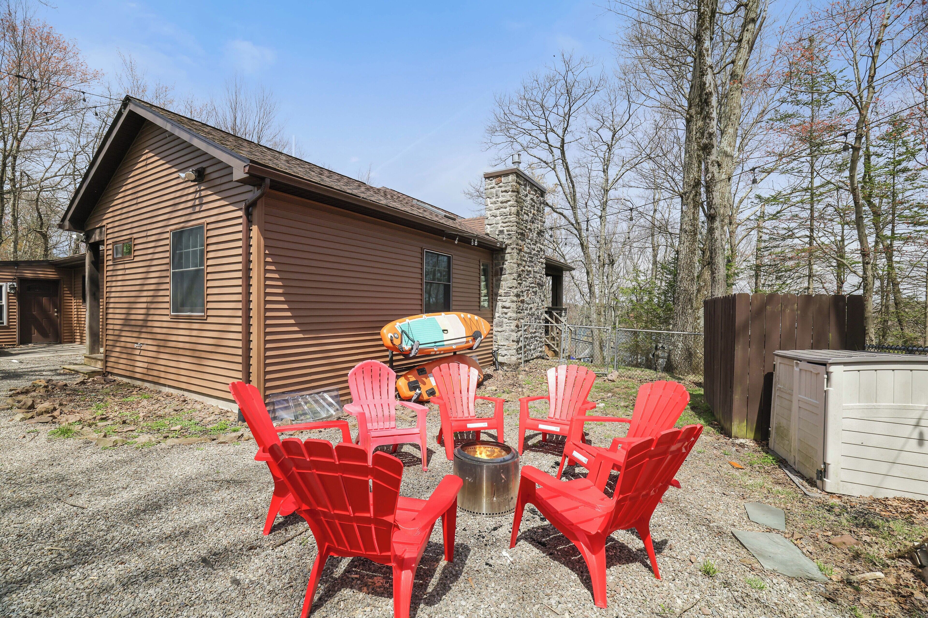 A wooden cabin with a stone chimney, surrounded by a gravel patio with red Adirondack chairs and a fire pit.