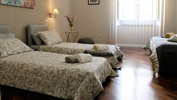 A hotel room with three beds, a small table with a vase of flowers, a wall-mounted lamp, and a window with blinds. - B&B Binario 39 (Naples)