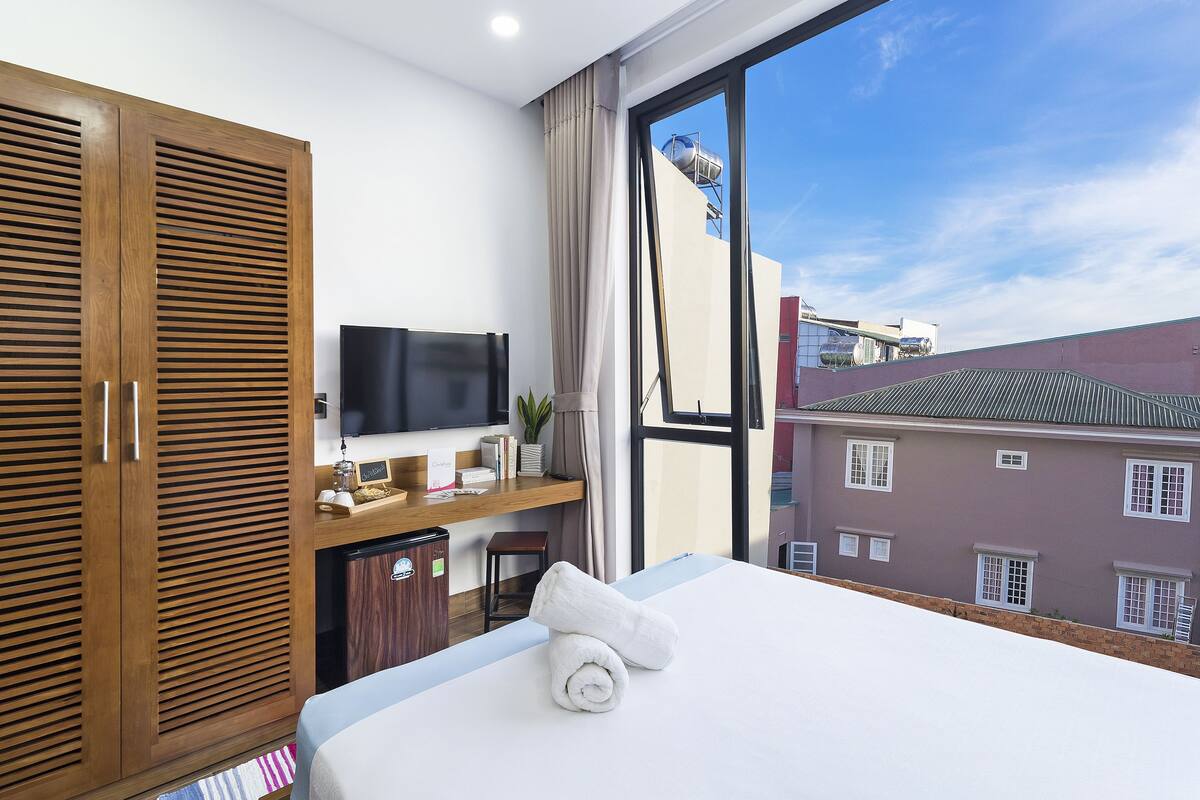A hotel room with a large bed, wooden wardrobe, TV, and a view of buildings through a large window.