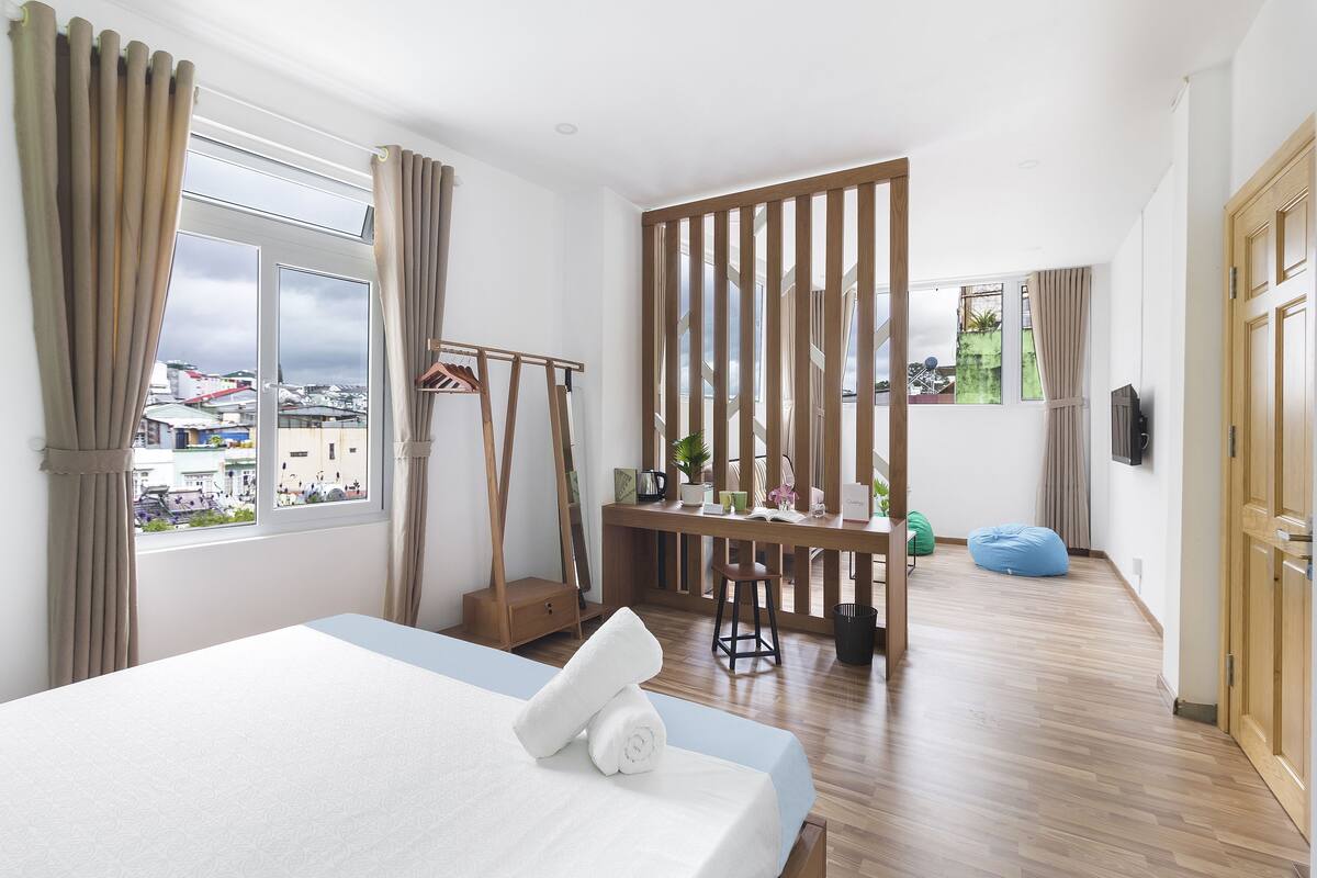 A hotel room with a bed, a desk, a chair, a wooden partition, and a view of the city.