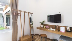 Habitación de hotel con un escritorio de madera, un televisor montado en la pared, una ventana con cortinas, un taburete pequeño y un cesto de basura.