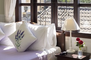 Un lit avec du linge de lit blanc et des oreillers lavande, une table de chevet avec une lampe et un vase de roses rouges, et une fenêtre ornée de barreaux en fer décoratifs.