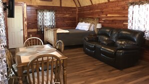 Un accogliente interno di una baita con soffitto in legno, un letto con cuscini, un divano in pelle, un tavolo da pranzo con sedie e una ventola a soffitto.