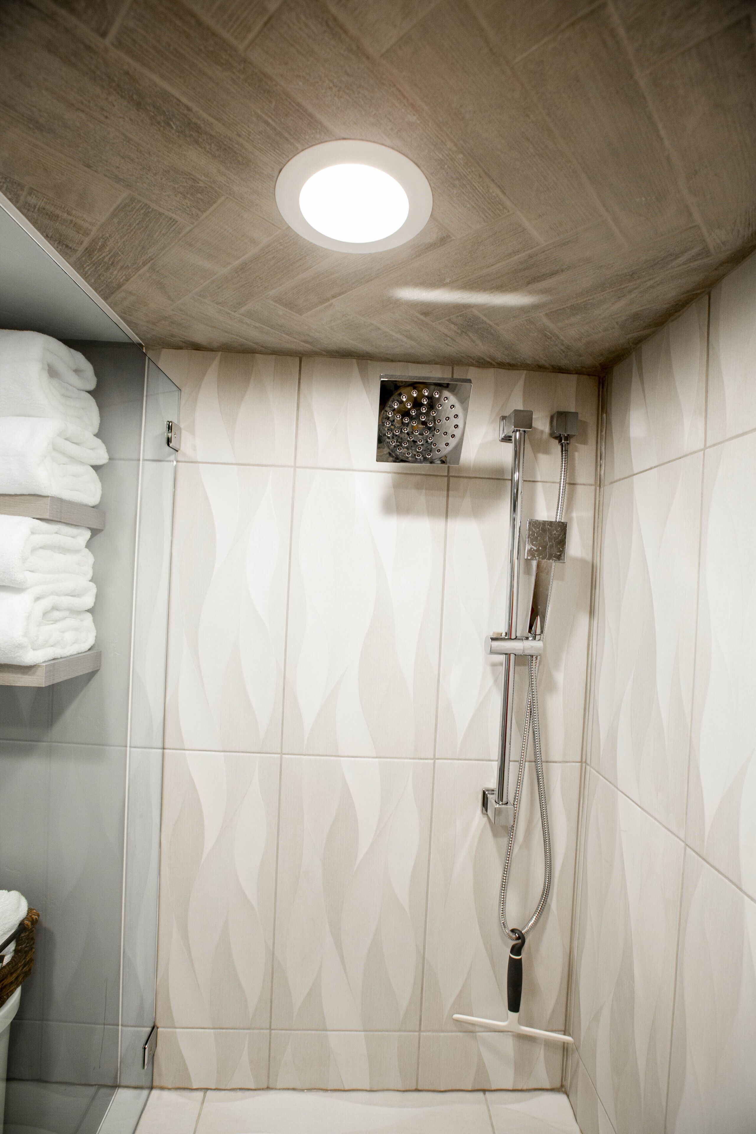 Een douche met handdouche en een muurkranen, betegelde wanden met een geometrisch patroon, en een plank met opgevouwen handdoeken.