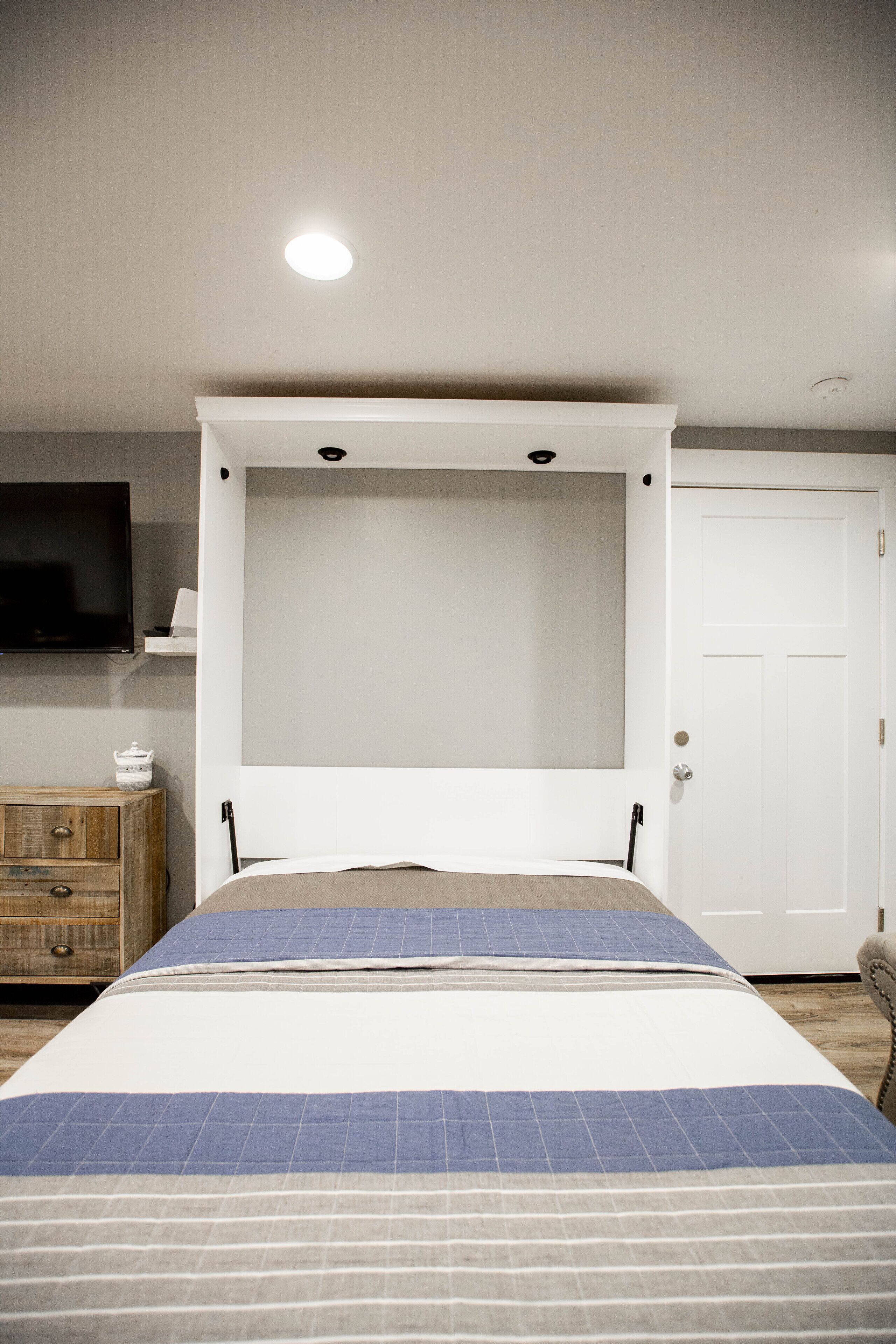 A bedroom with a bed, a television on the wall, a wooden dresser, and a white door.