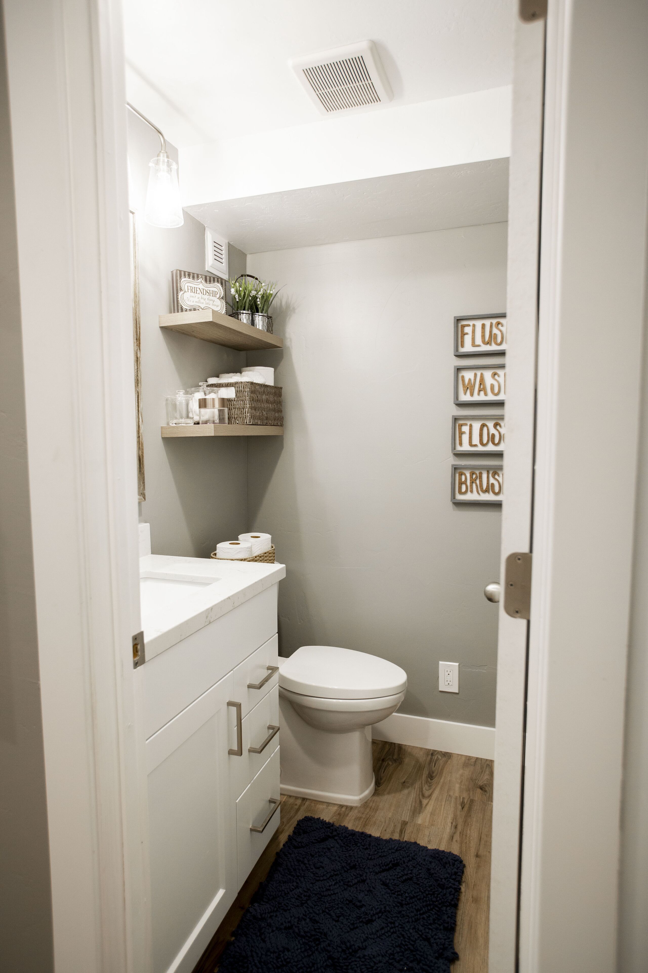 Une petite salle de bain propre, avec des toilettes, des meubles blancs, une étagère contenant des articles de toilette et un tapis bleu foncé.