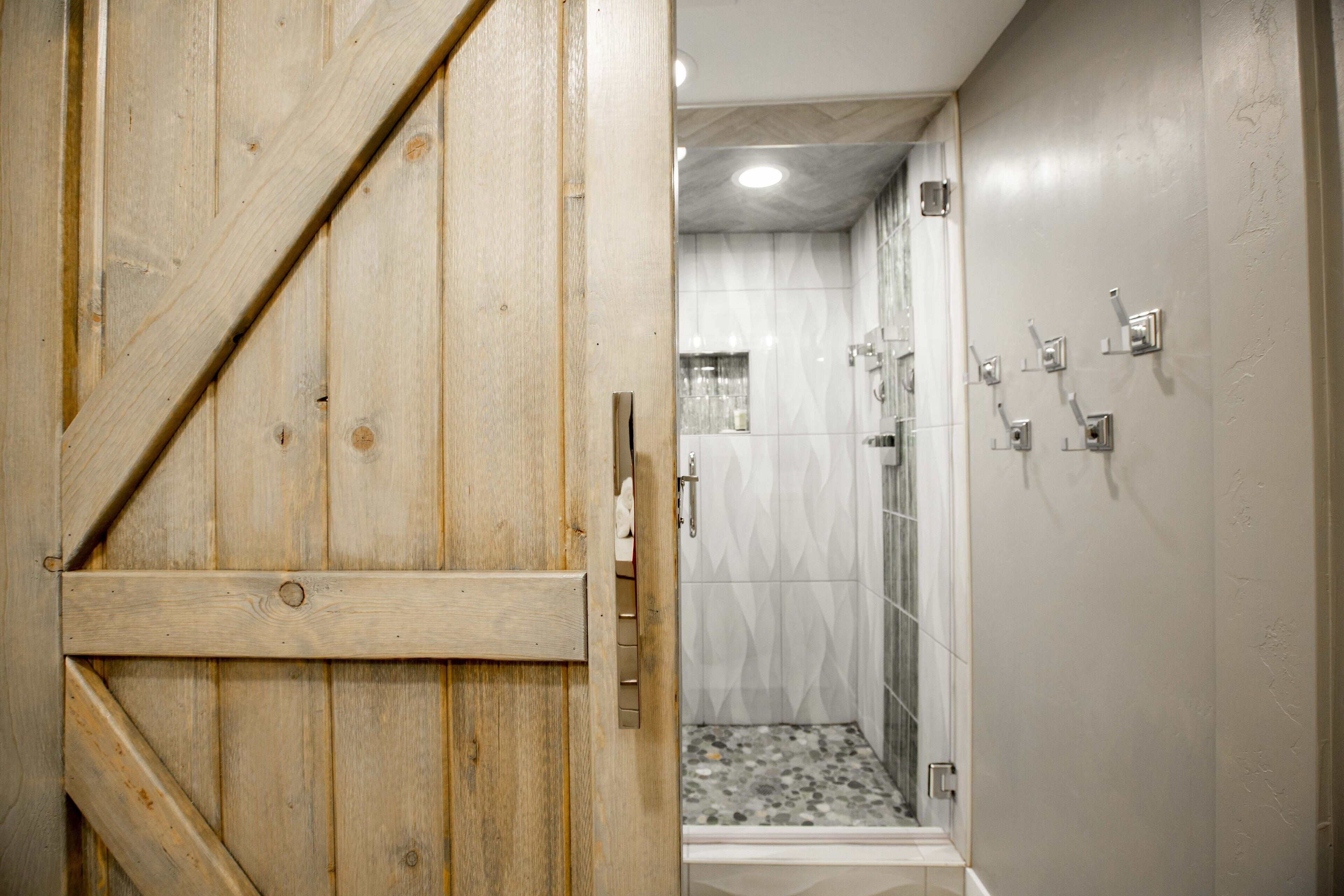 A wooden door leading to a bathroom with a shower, tiled walls, and a tiled floor.