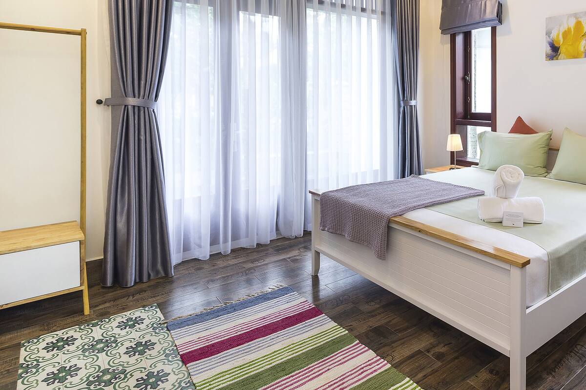 A neatly arranged bedroom with a bed, bedside tables, a nightstand, a window with curtains, and a wall-mounted artwork.