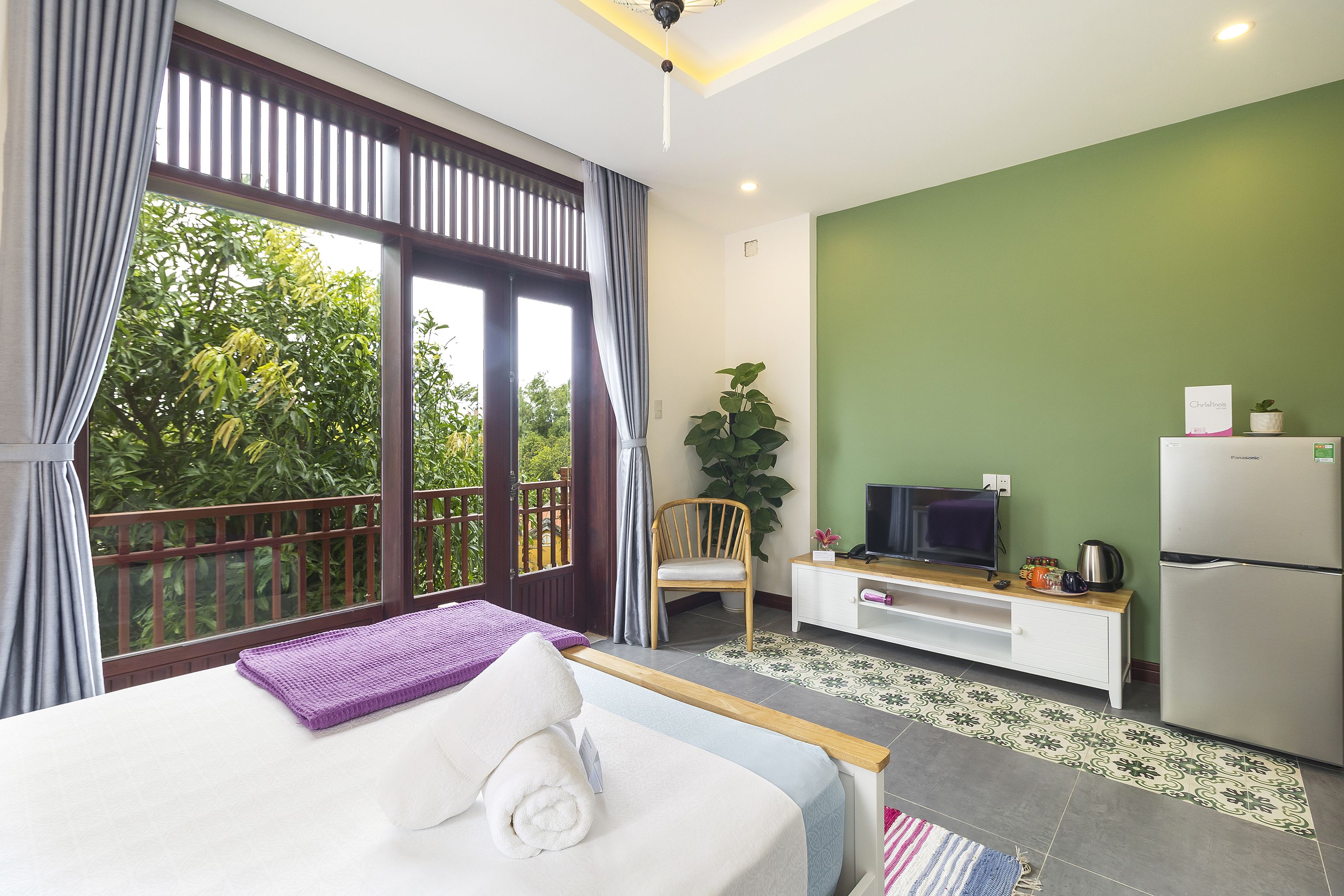 Un dormitorio con cama, televisión, refrigerador y balcón con vistas a la vegetación.