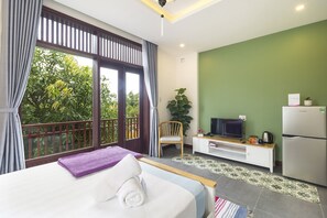 A bedroom with a bed, a TV, a refrigerator, and a balcony with a view of greenery. - Christina's Hoi An - Tra Que Oasis (Da Nang)