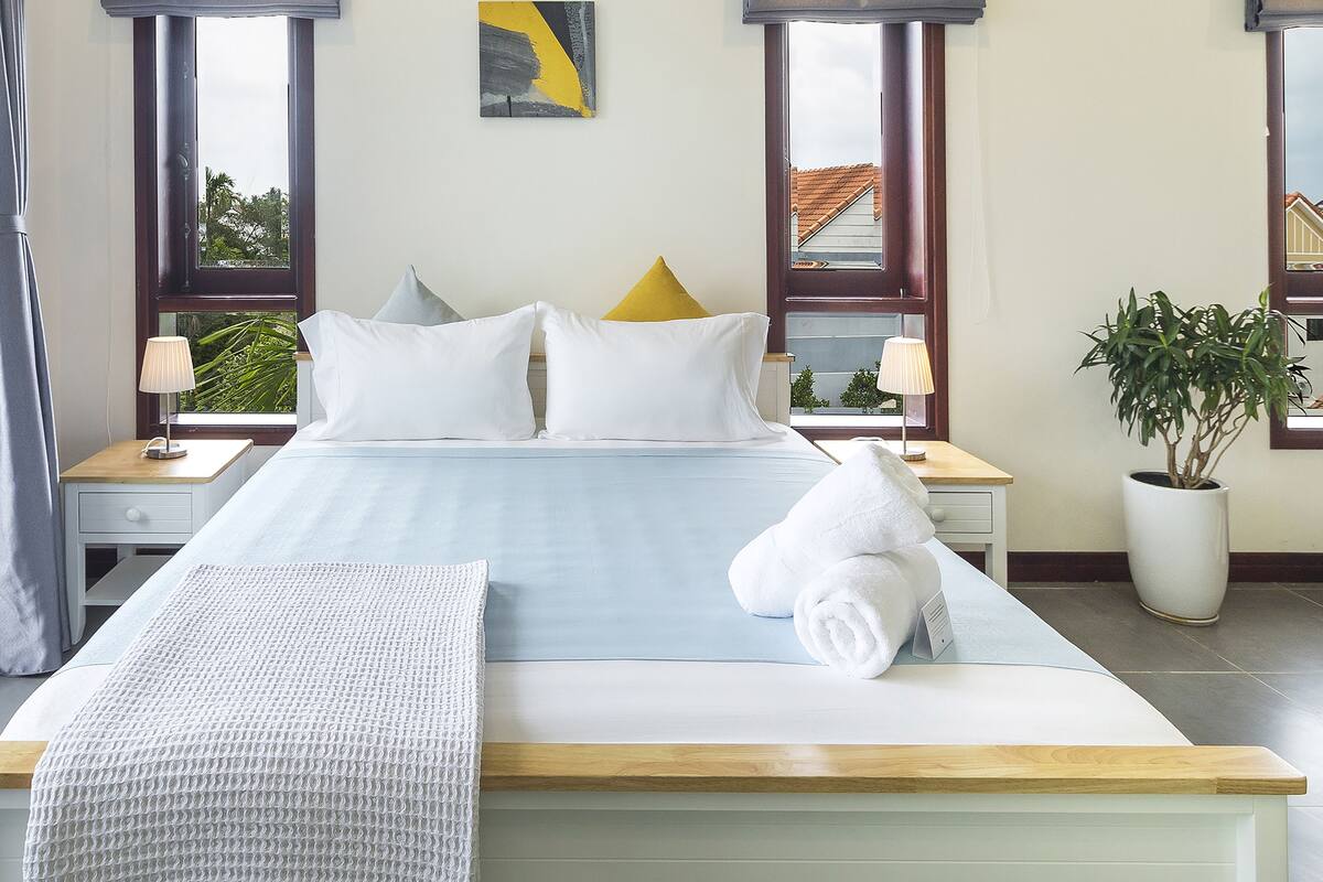 A neatly made bed with white linens and pillows, a bedside table with a lamp, and a large window with a view of the outside.