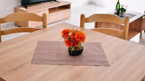 Une table à manger en bois, sur laquelle est posé un vase rempli de fleurs oranges, est entourée de chaises en bois. Un téléviseur est fixé au mur au fond de la pièce.