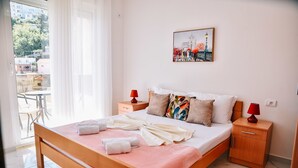 Un lit bien fait, avec des oreillers et une couverture, une table de chevet avec une lampe, et une fenêtre donnant sur des bâtiments et des arbres.