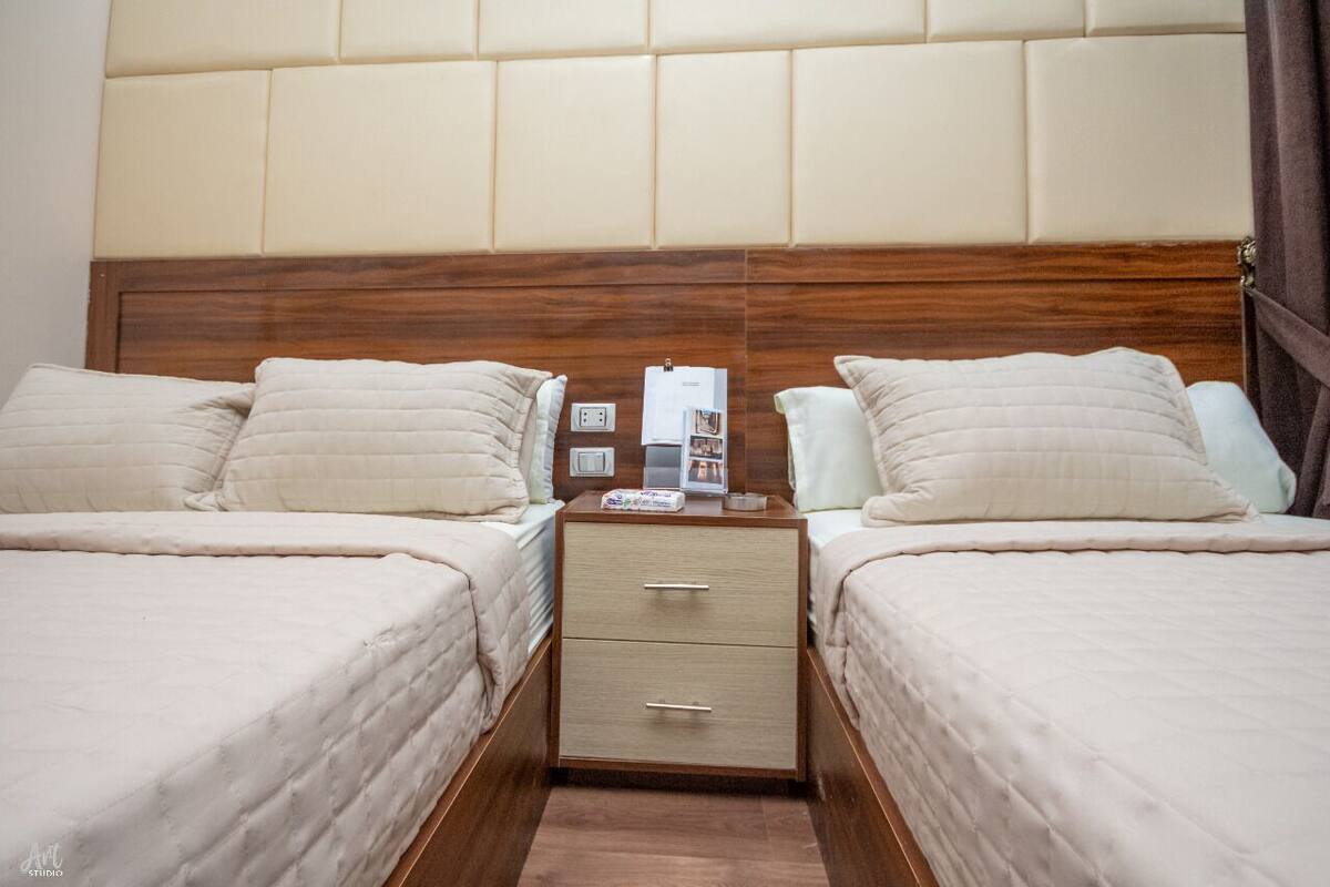 Habitación de hotel con dos camas, cabecero de madera, mesitas de noche con lámparas y un aparador con teléfono y revista.