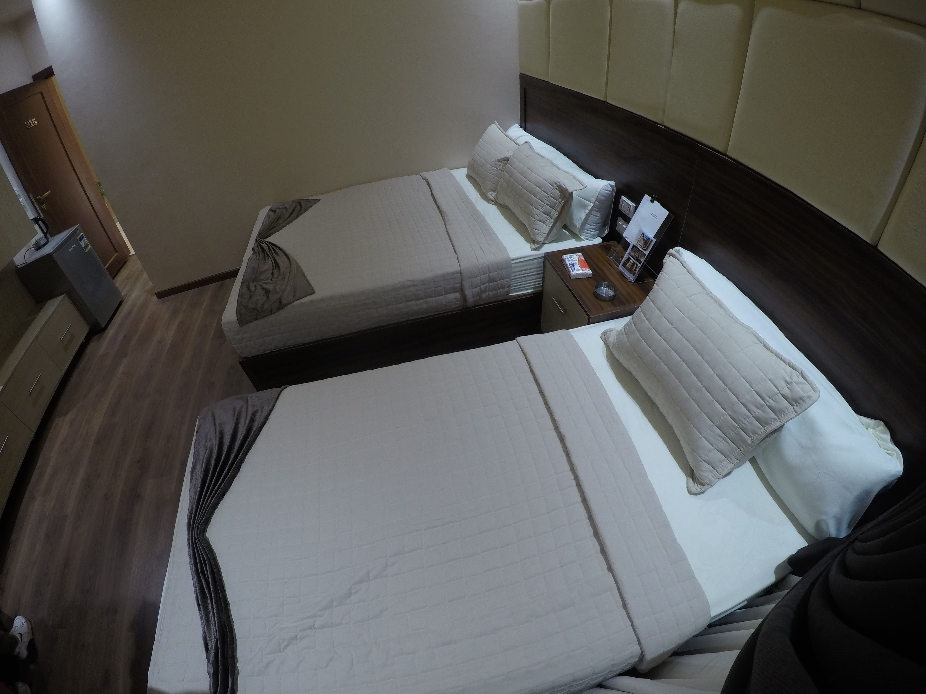 A hotel room with two beds, a wooden wardrobe, a desk, and a nightstand with a lamp and a magazine.
