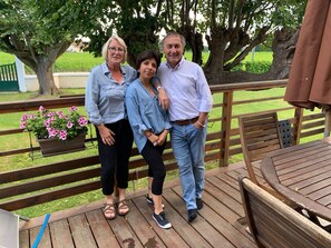 A wooden deck with a table and chairs, surrounded by greenery and a large tree. - Renovated 3 épis barn with 1 to 2 air-conditioned bedrooms for 2 to 6 people. (ASTIS)