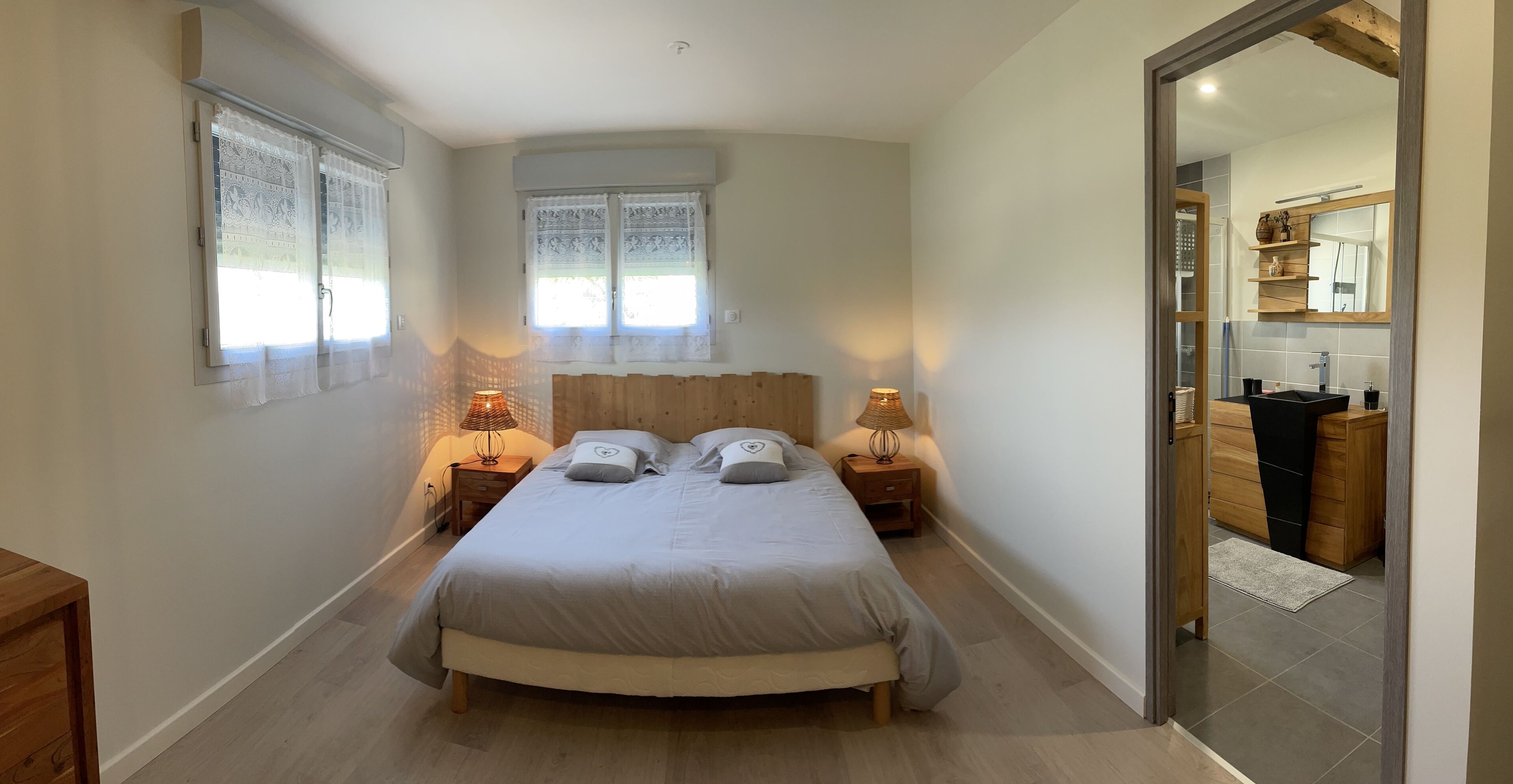 Une chambre à coucher avec un lit, deux tables de chevet, une fenêtre ornée de rideaux en dentelle et une tête de lit en bois.