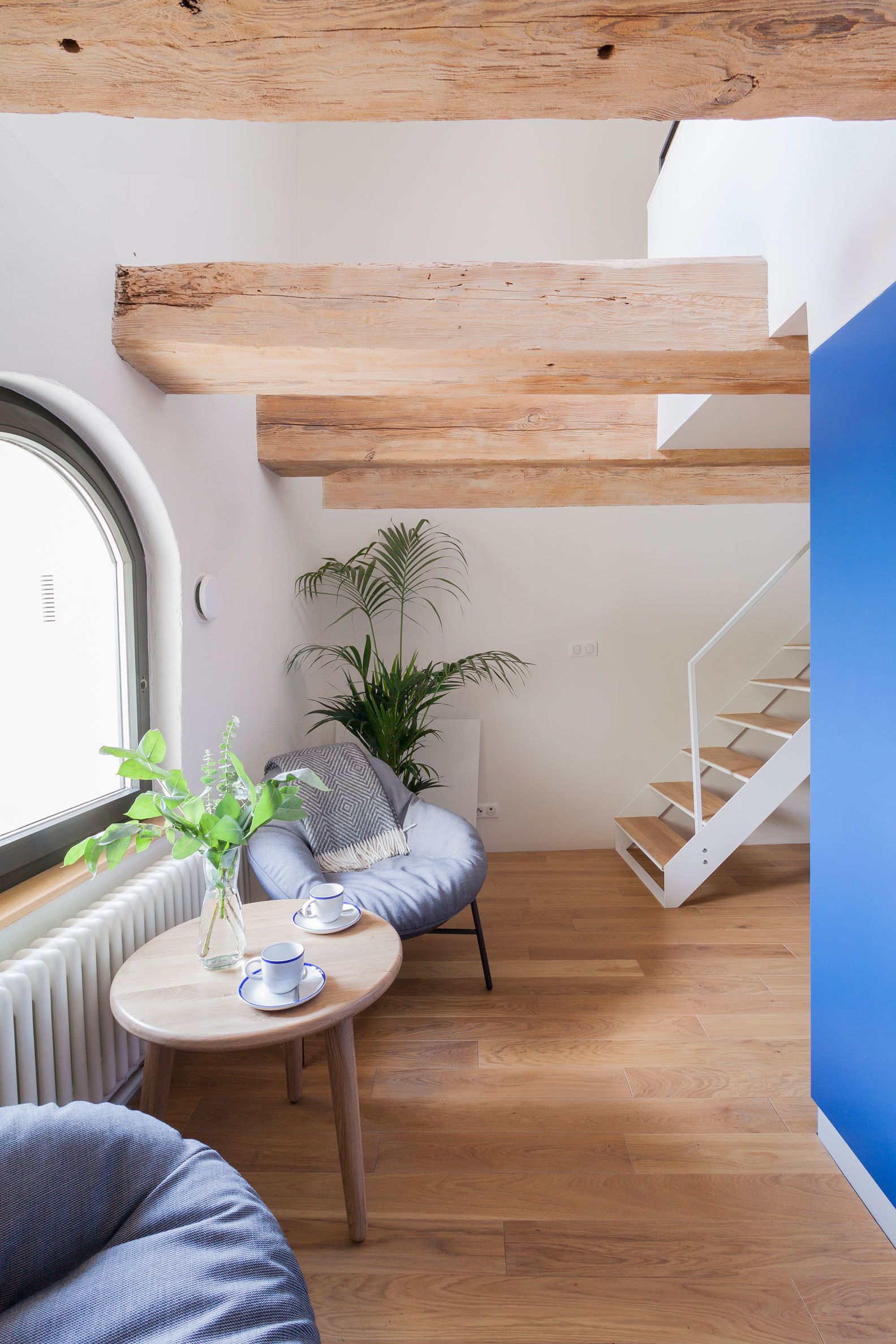 A modern living room with a wooden floor, a round wooden table with a vase and cups, a blue sofa, a staircase, and a large window.
