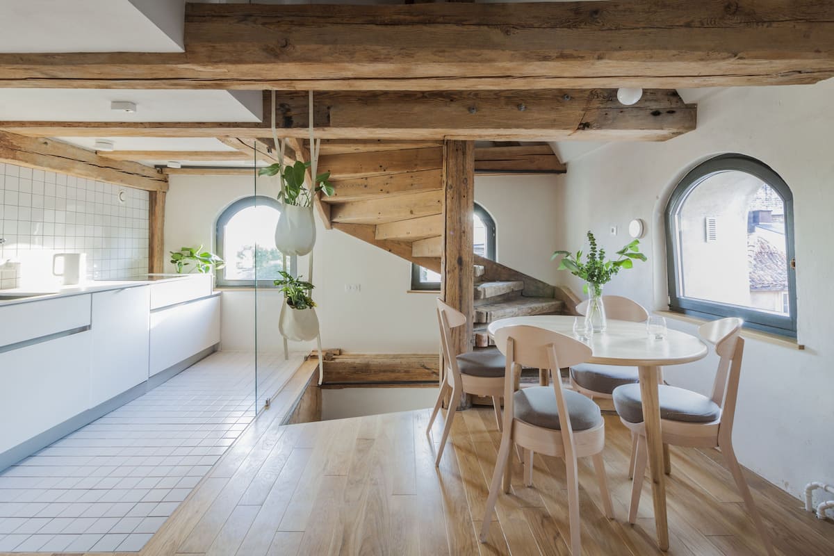 A modern kitchen with wooden beams, a dining area with a round table and chairs, and a view through a window.