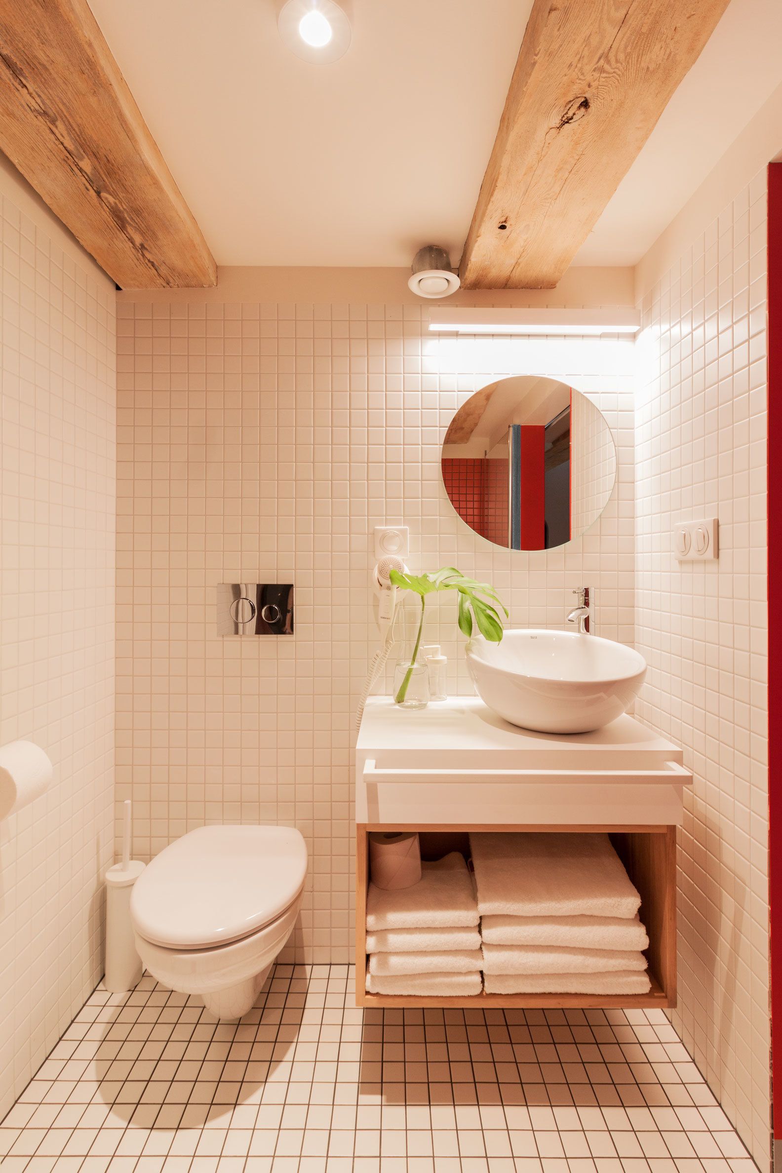 Un bagno con pavimento in piastrelle bianche, un WC bianco, un lavandino con uno specchio sopra e uno scaffale in legno con asciugamani.