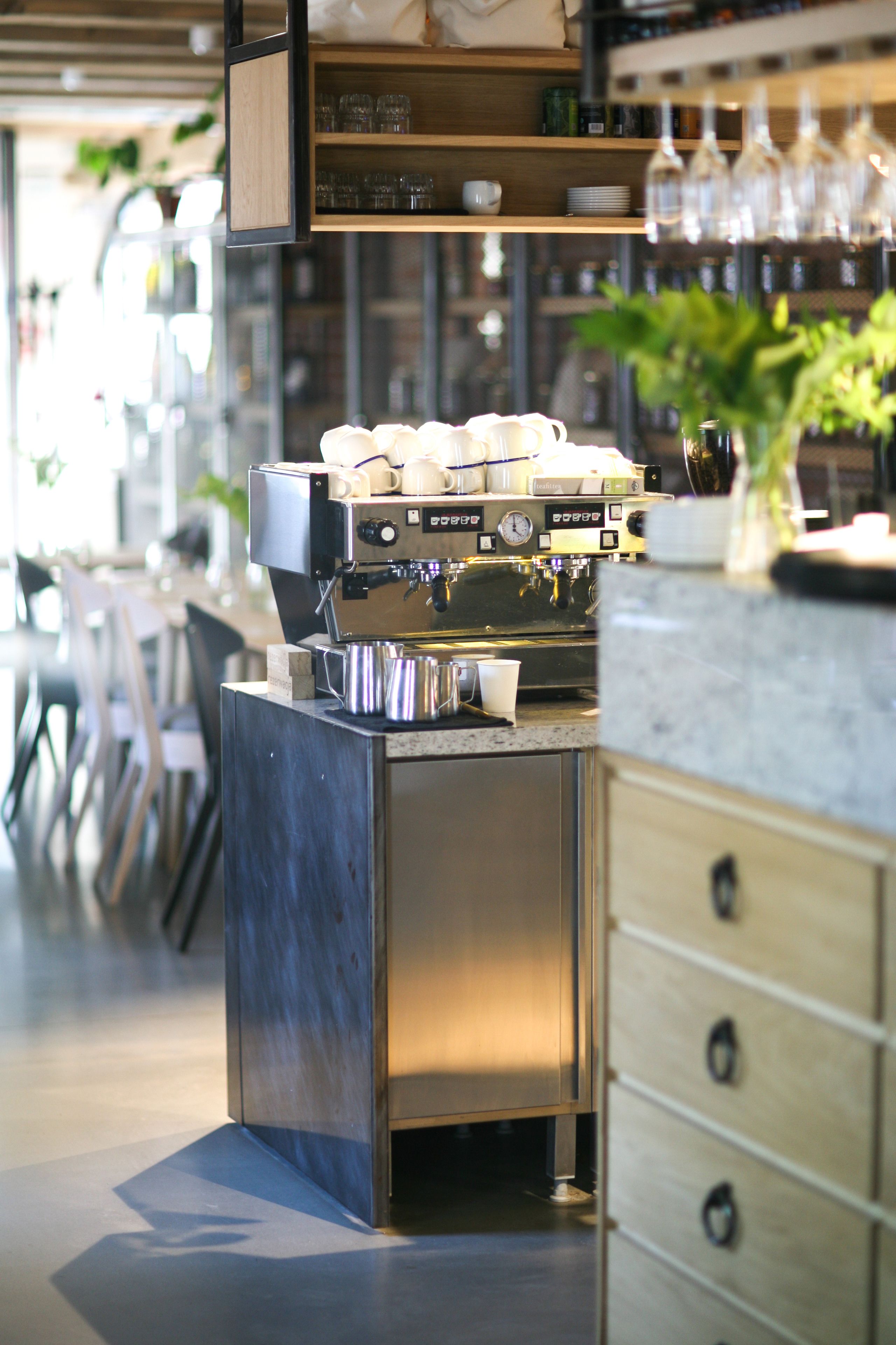 A coffee machine with multiple portafilters, a stainless steel counter, and a wooden shelf with glassware.
