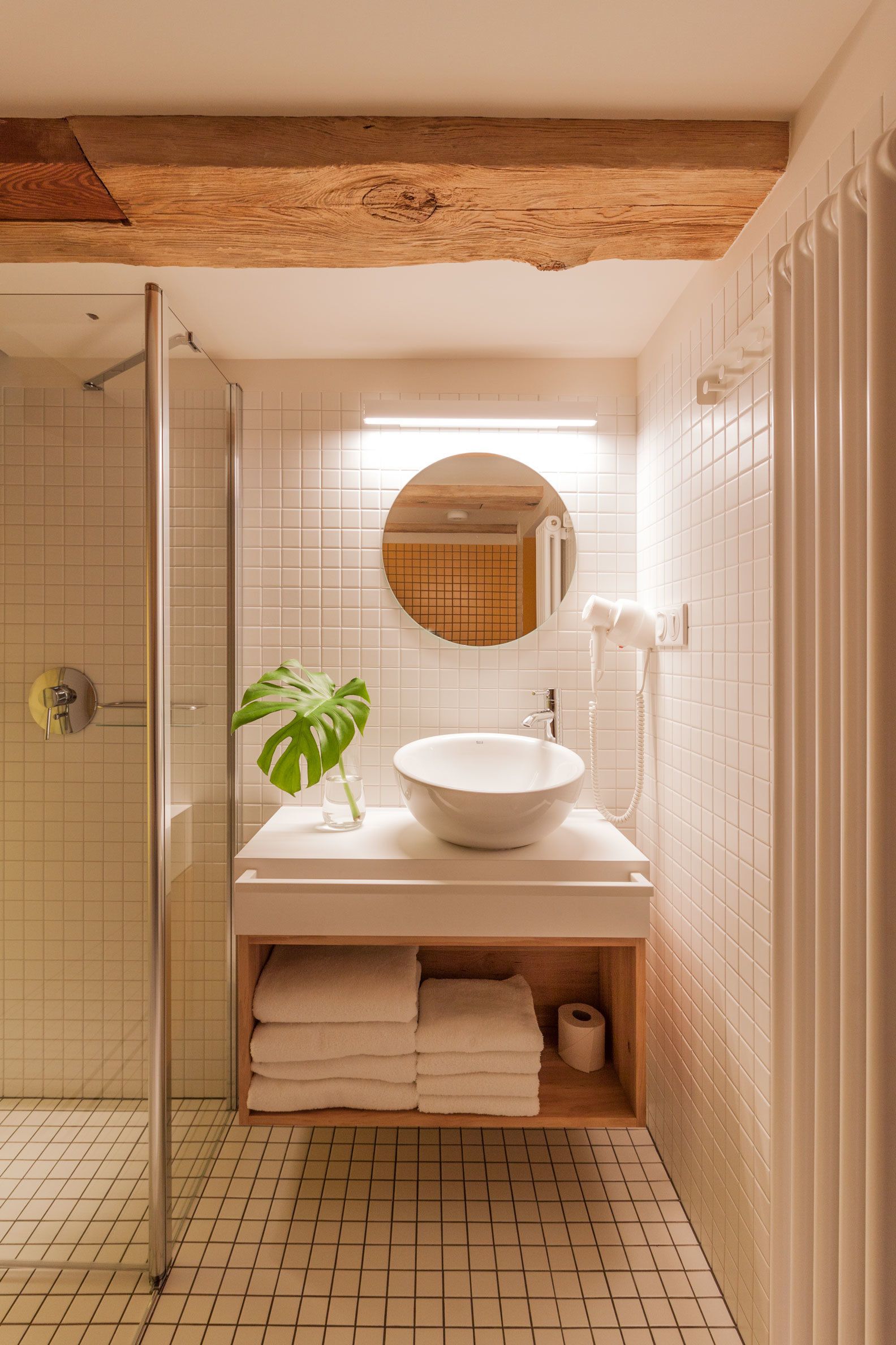 Une salle de bain moderne avec une douche à parois de verre, un miroir rond, un lavabo blanc et une étagère en bois où sont posées des serviettes.