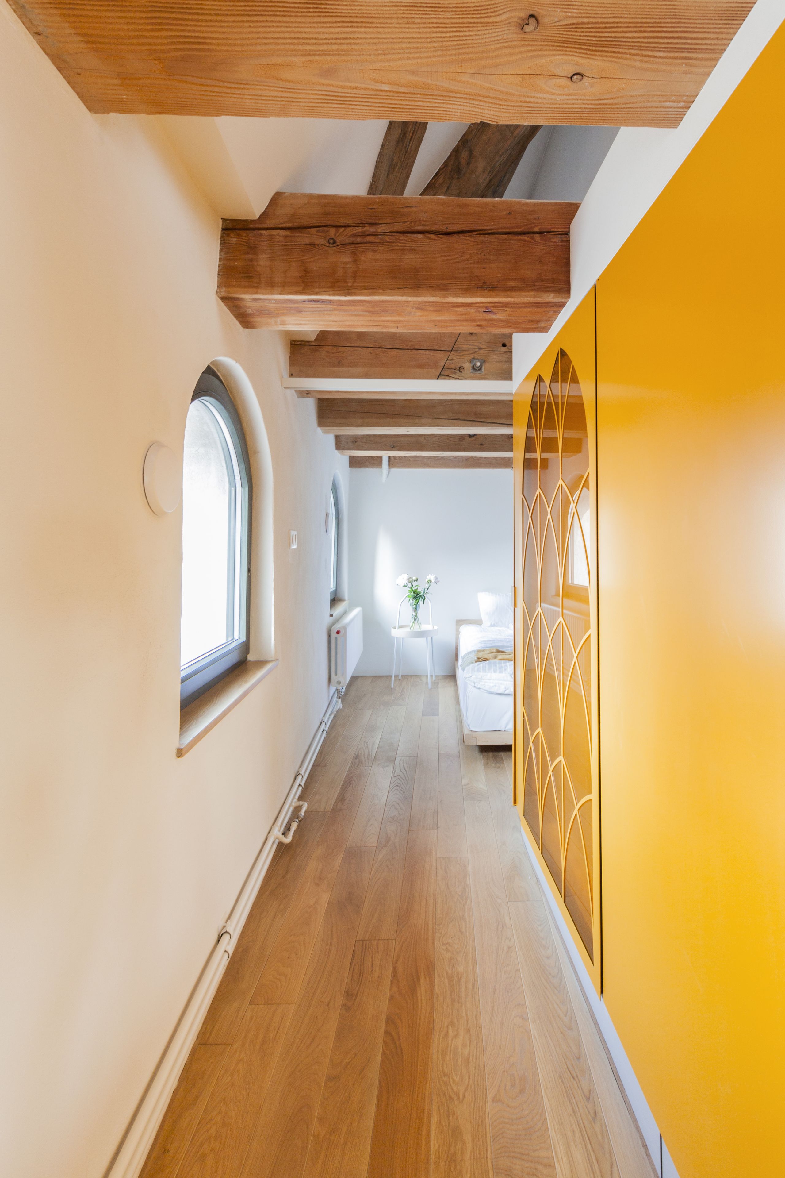 Un pasillo con vigas de madera, una pared amarilla y una cama al fondo.