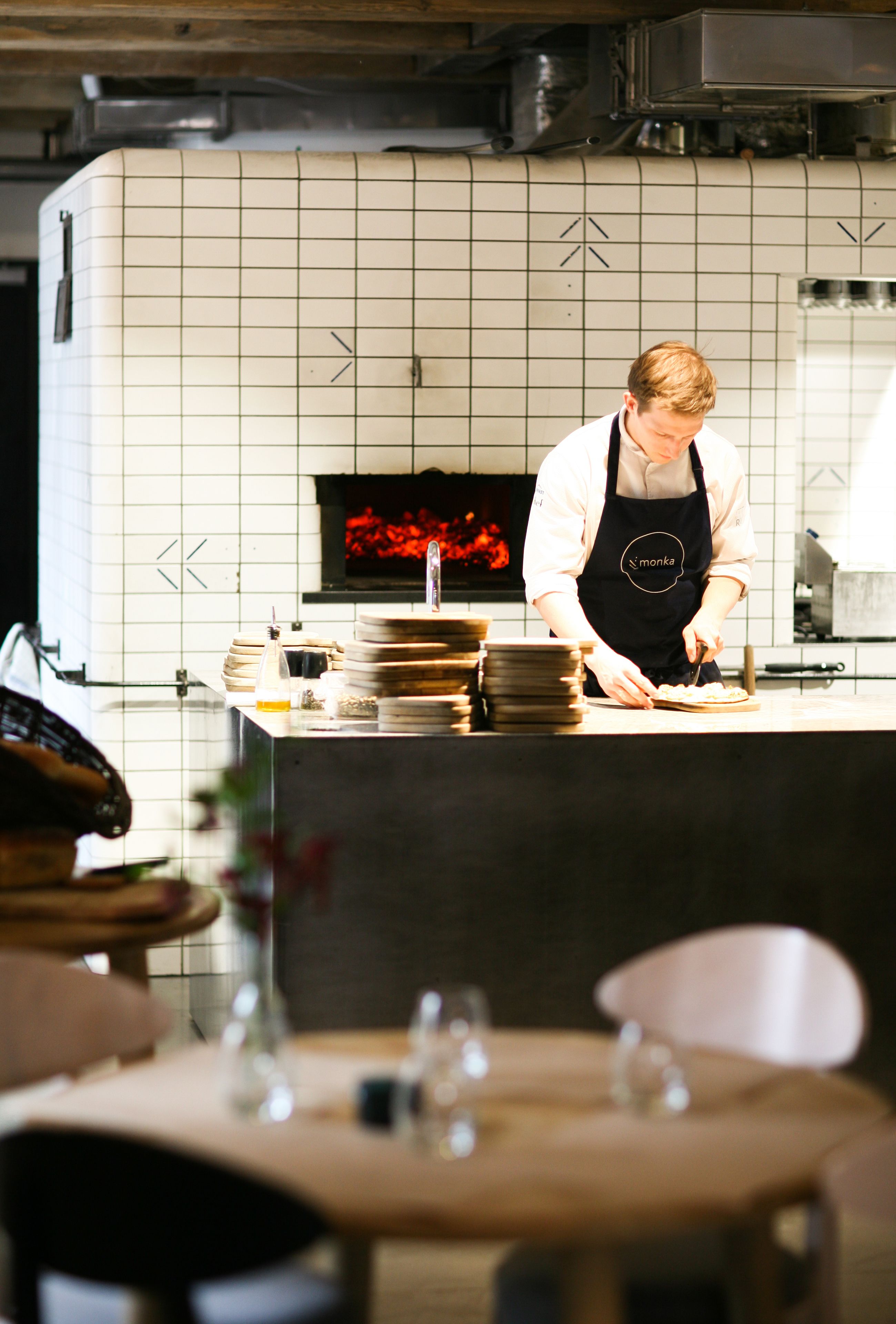 Un chef en una cocina con salpicadero de azulejos, un horno de pizza y una encimera con platos apilados y una botella.
