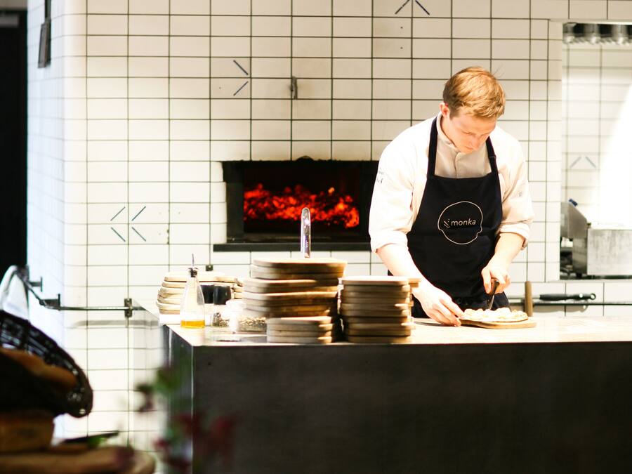 A chef in a kitchen with a tiled backsplash, a pizza oven, and a counter with stacked plates and a bottle.