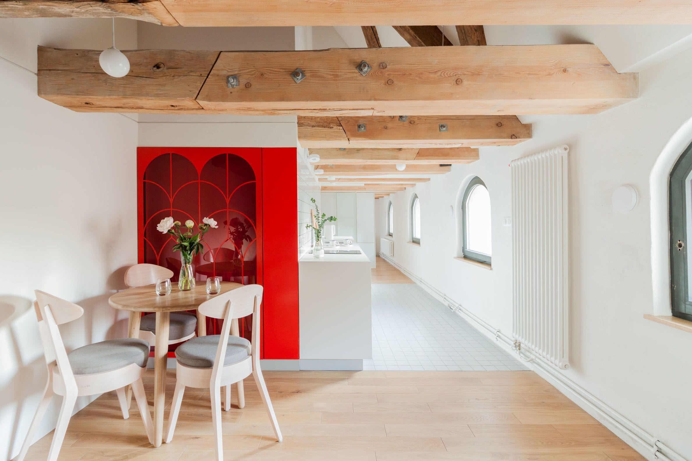 A modern dining area with wooden beams, a red accent wall, and a view of a hallway with arched windows.