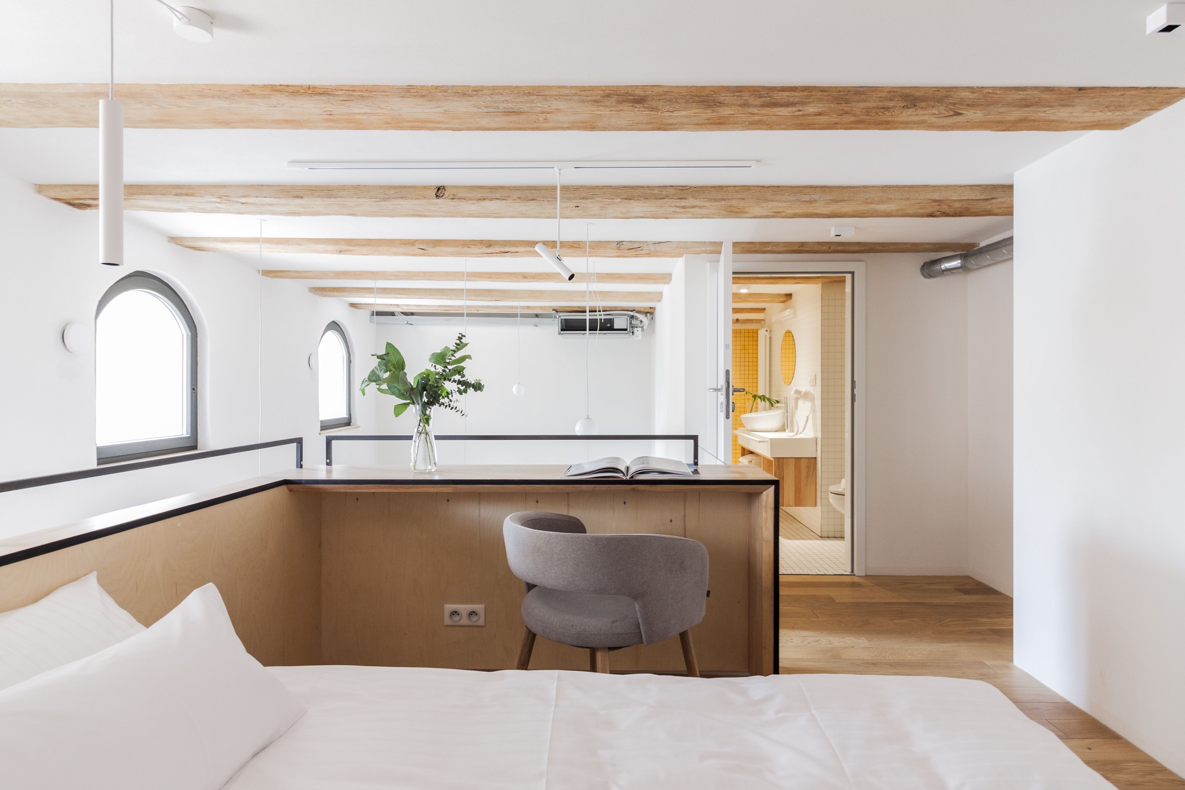 A modern hotel room with a wooden ceiling, a bed with white linens, a grey chair, and a small desk with a plant.