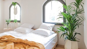 Un dormitorio con cama, ropa de cama blanca, suelo de madera, una planta grande en maceta y una planta colgante.