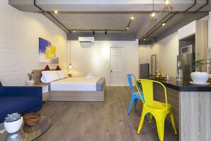 A modern hotel room with a bed, a sofa, a dining area with a chair, and a kitchen area with a refrigerator.