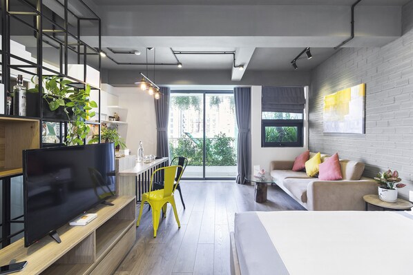 A modern living room with a flat-screen TV, a yellow chair, a sofa with pink and yellow cushions, a small round table with a plant, and a large window with a view of buildings and trees.