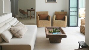 Ein geräumiges Wohnzimmer mit hoher, gewölbter Decke, einem weißen Sofa mit Kissen, zwei Korbstühlen mit braunen Kissen, einem Holztisch und einem gerahmten Bild an der Wand.