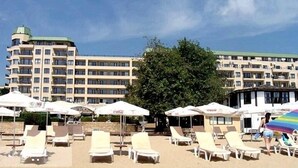 Een hotel met meerdere verdiepingen, terras met parasols en een grote boom op de voorgrond.
