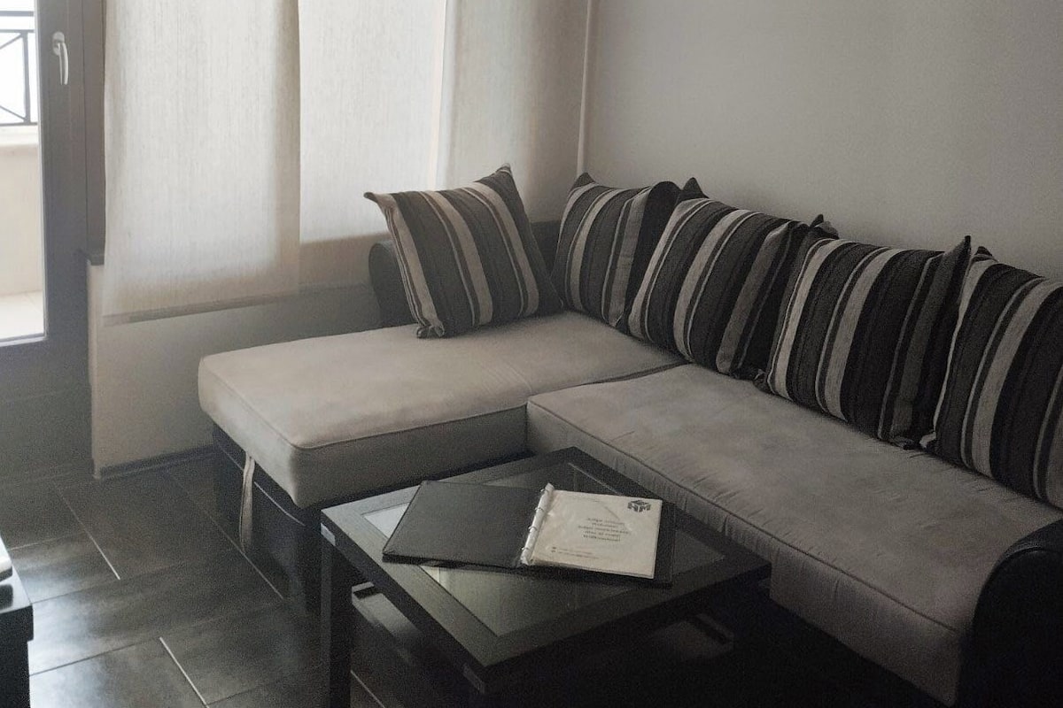 A corner living room with a grey sofa, striped cushions, a small table, and an air conditioning unit on the wall.