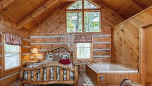 A log cabin bedroom with a wooden bed, a small bedside table, a nightstand, a window with curtains, a large window with a view of trees, and a wooden bathtub. - View! Private Studio Cabin,Hot Tub,RELAX,Honeymoon (Sevierville)