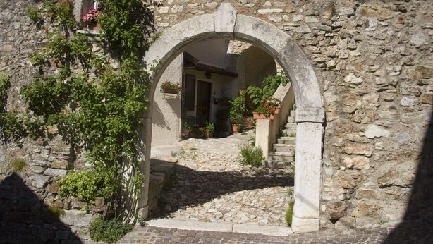 Un portail en pierre en forme d’arc donne accès à une cour intérieure, où un chemin pavé serpente entre des murs de pierre et des plantes en pot.