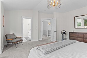 A bedroom with a bed, a chair, a dresser, a mirror, and a hallway leading to a staircase.