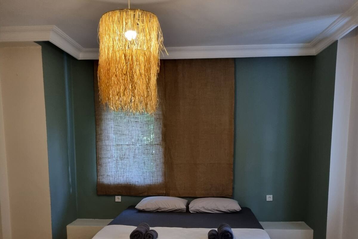 A bedroom with a large bed, a hanging light fixture, and a herringbone-patterned wooden floor.