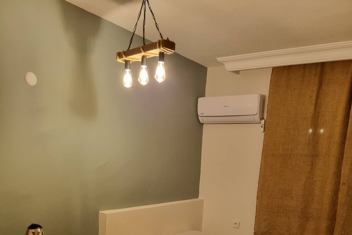 A small, well-lit room with a bed, a small desk, a kettle, and a heater.
