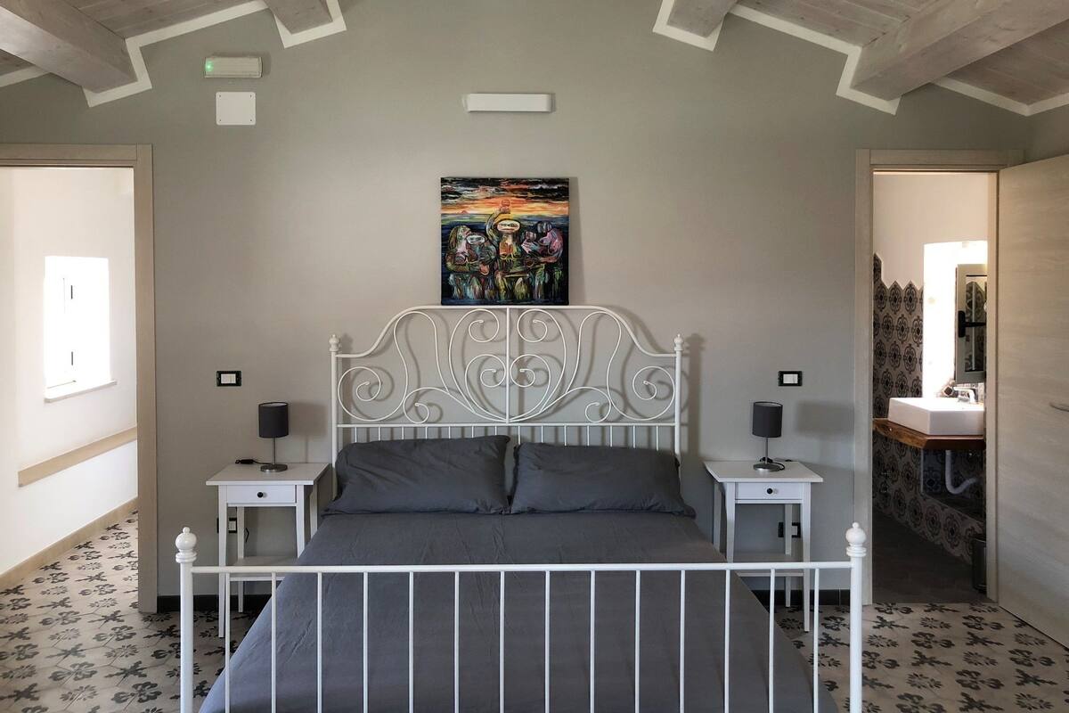 Une chambre avec un lit en métal blanc, deux tables de chevet avec des lampes, un tableau au mur et une salle de bain visible à travers une porte ouverte.