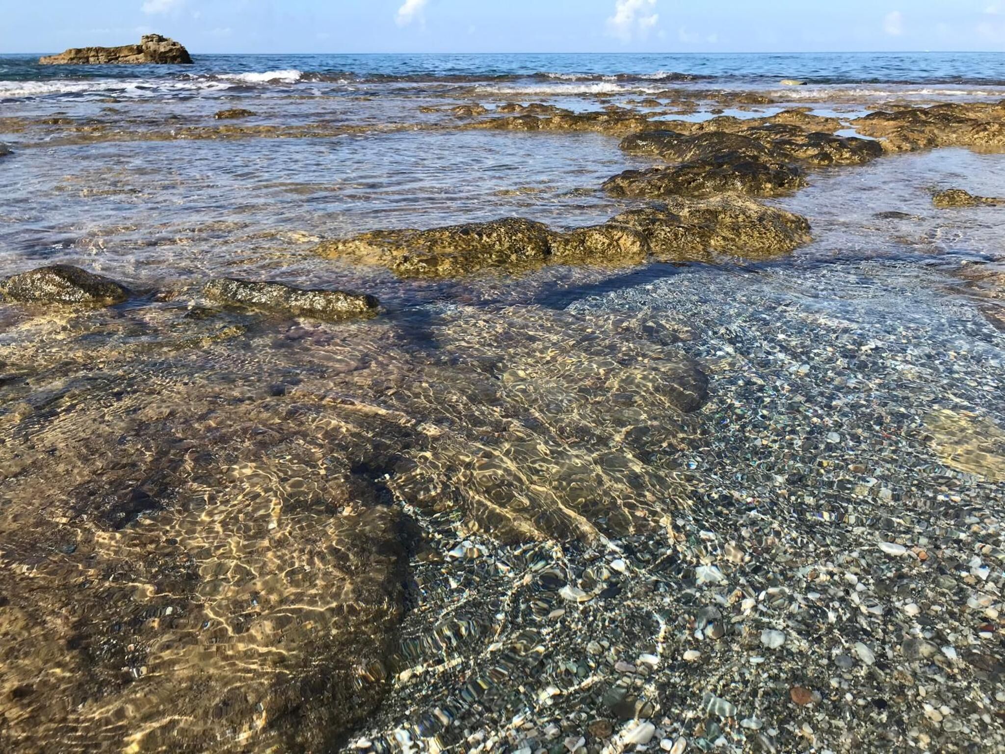 A rocky shoreline with clear water, pebbles, and a distant island.