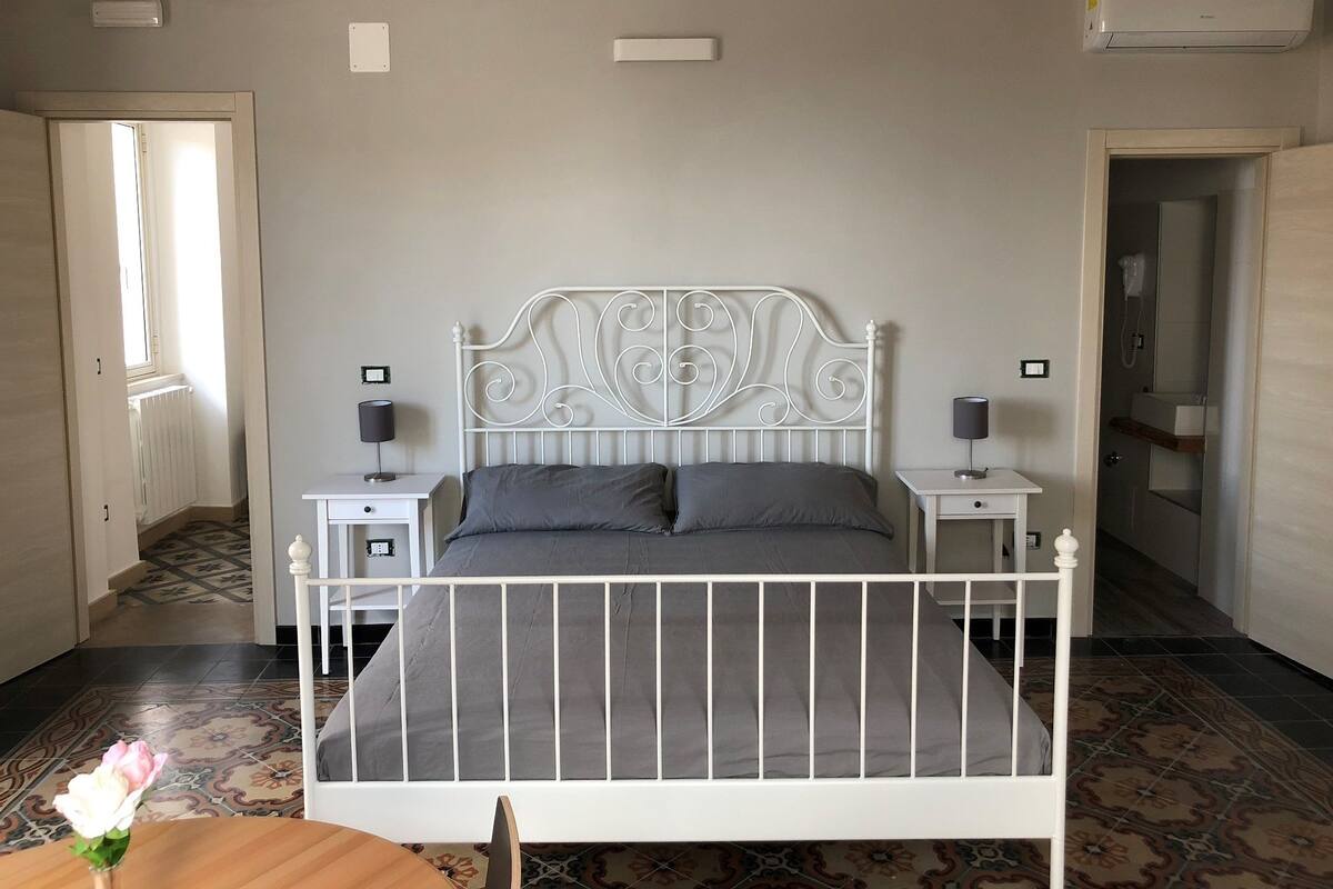 Une chambre à coucher avec un lit en métal blanc, une literie grise, deux tables de chevet avec des lampes, un radiateur et un sol à motifs.