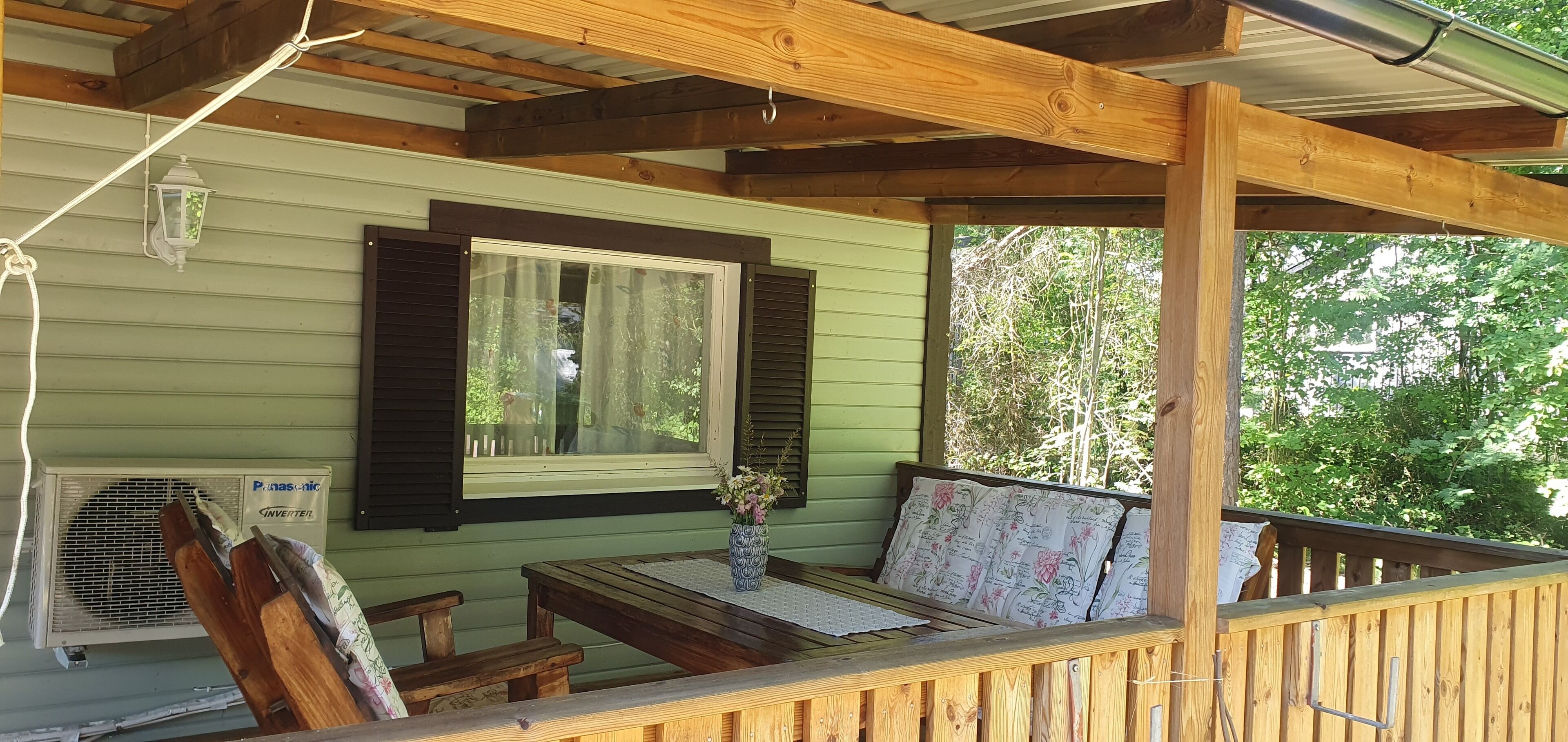 A covered porch with a wooden bench, a table with a vase of flowers, and a window with shutters.